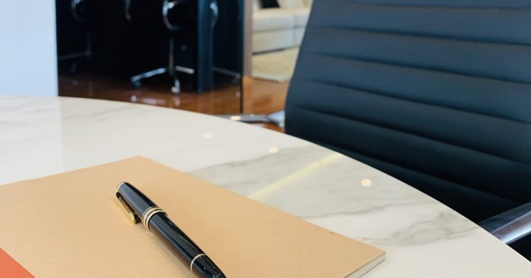 Cuaderno cerrado con una pluma encima sobre una mesa de juntas, con una silla vacía al lado